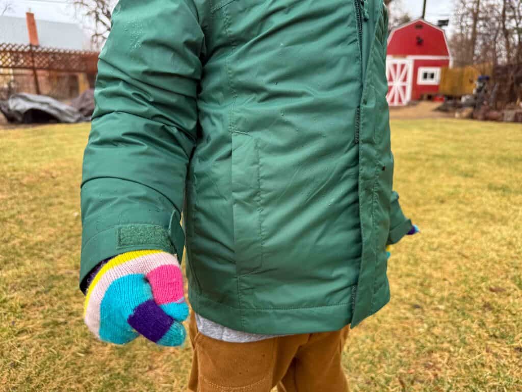 Child in green jacket with colorful mittens in backyard