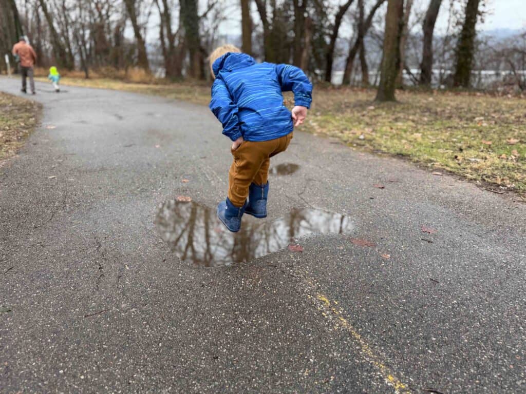 Young child jumping over puddle on park path in winter