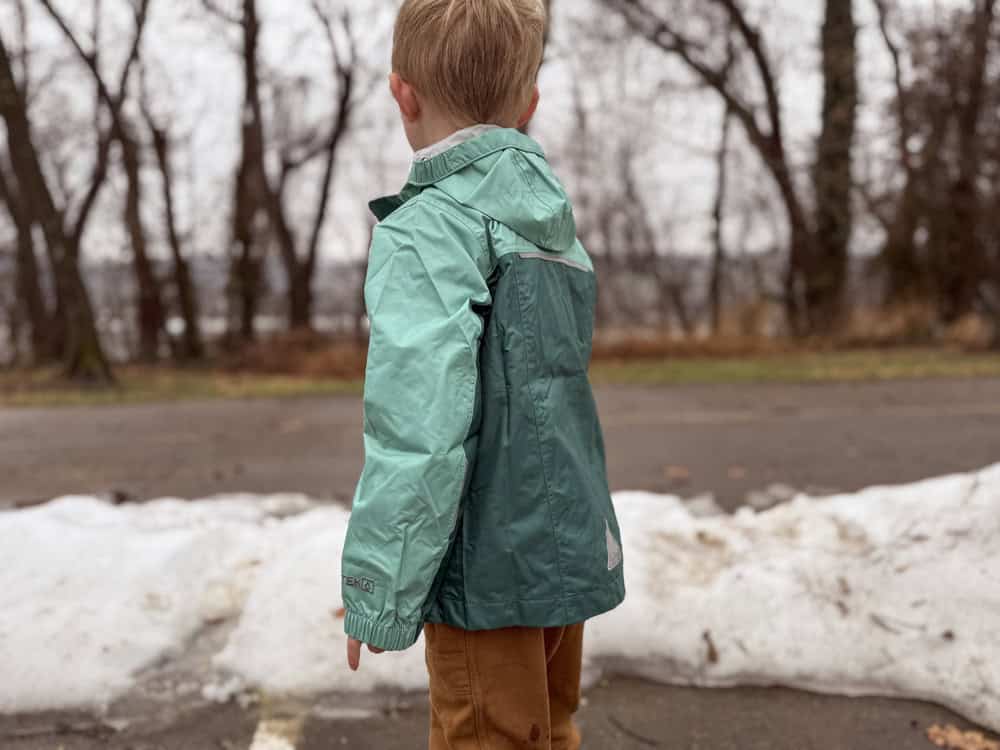 boy stands in front of snow wearing green two tone rain jacket