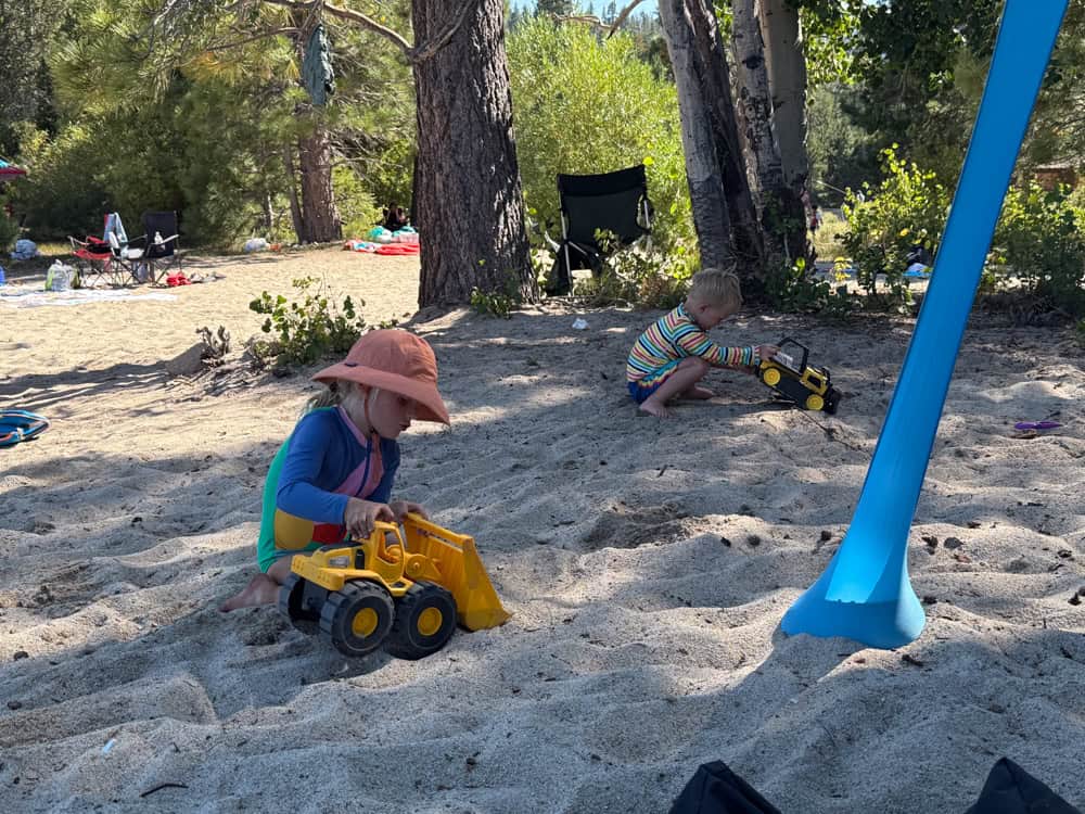 Two young children playing with toy trucks on sandy beach