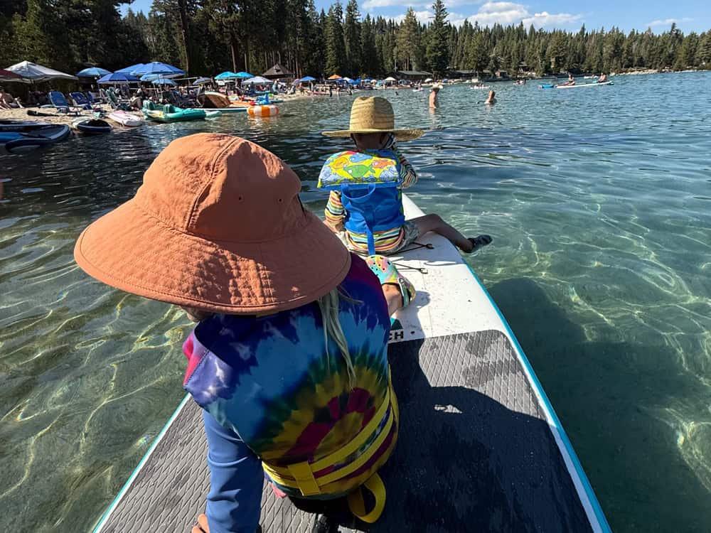 Two children sitting on paddleboard at busy mountain lake beach
