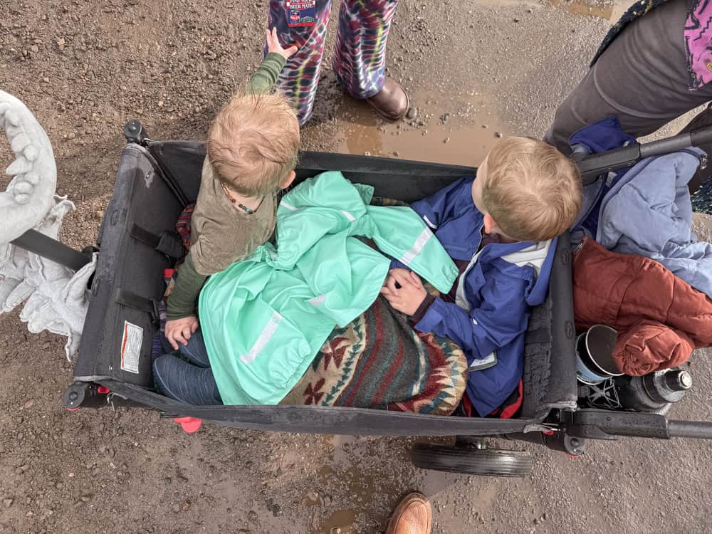 two kids sit in a wagon with a bunch of coats around them in the rain