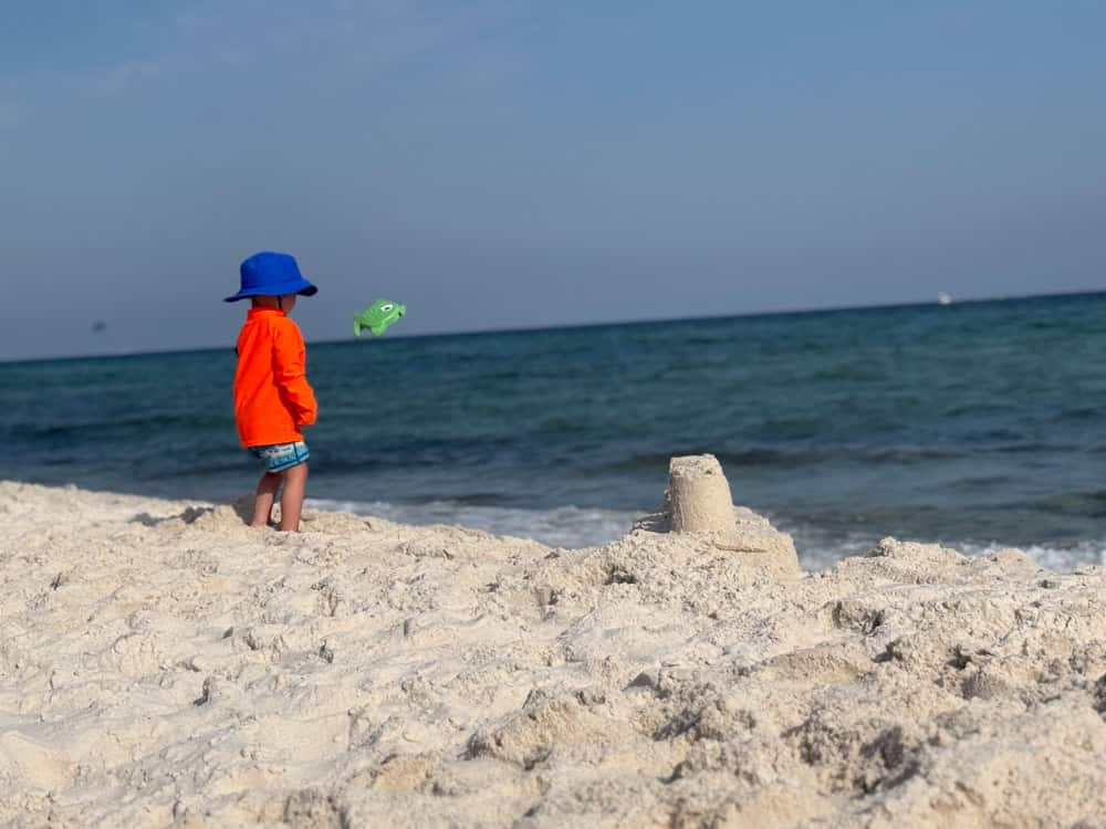 Young child in orange shirt gazing at ocean near sandcastle