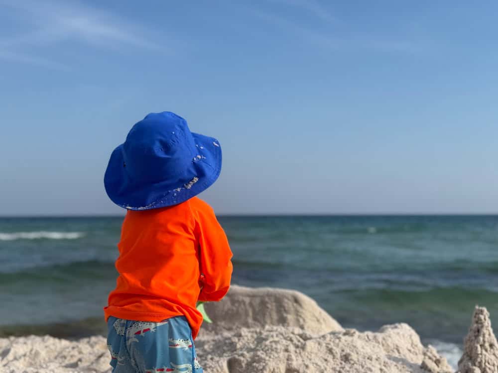Toddler in blue hat gazing at ocean by sandcastle