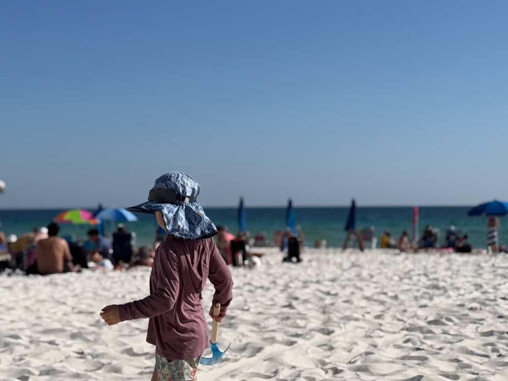 Child with sun hat and shovel walking on crowded beach