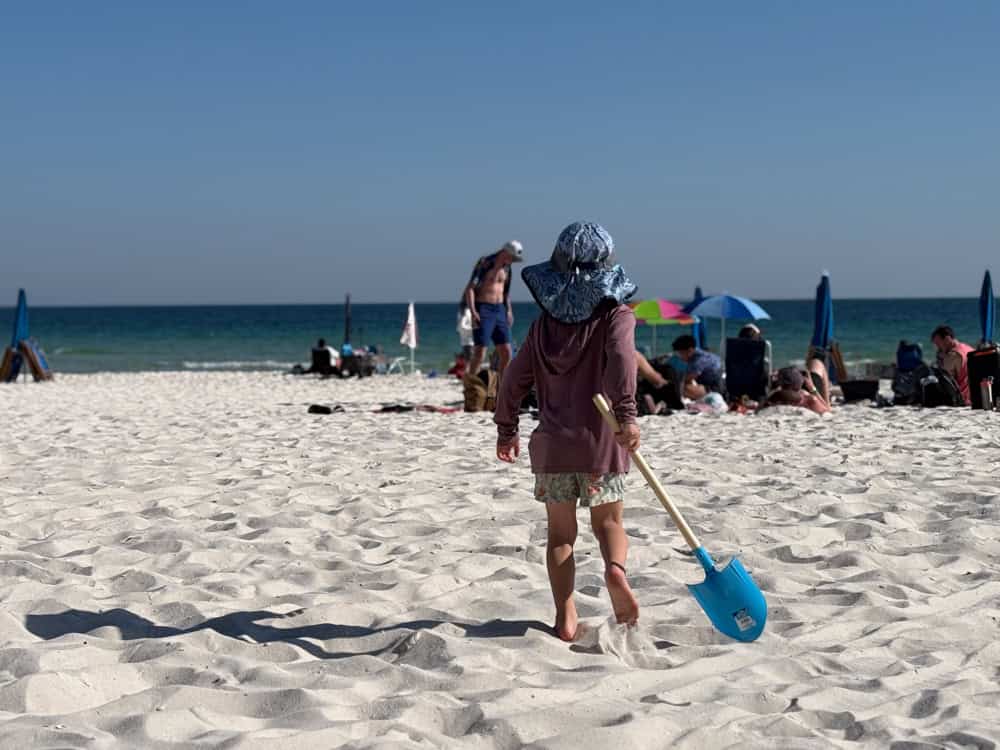 Child with blue shovel walking on white sandy beach