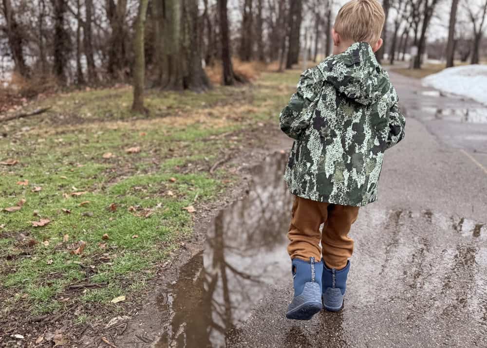 Young boy in camo jacket and rain boots near puddle