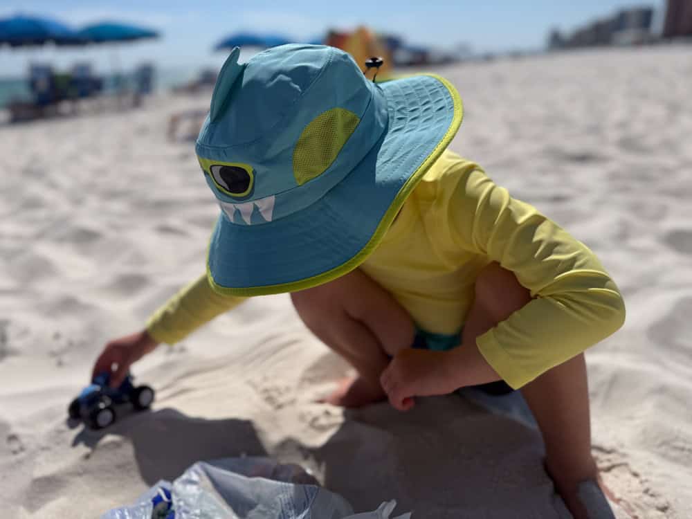Toddler in monster hat playing with toy car on beach