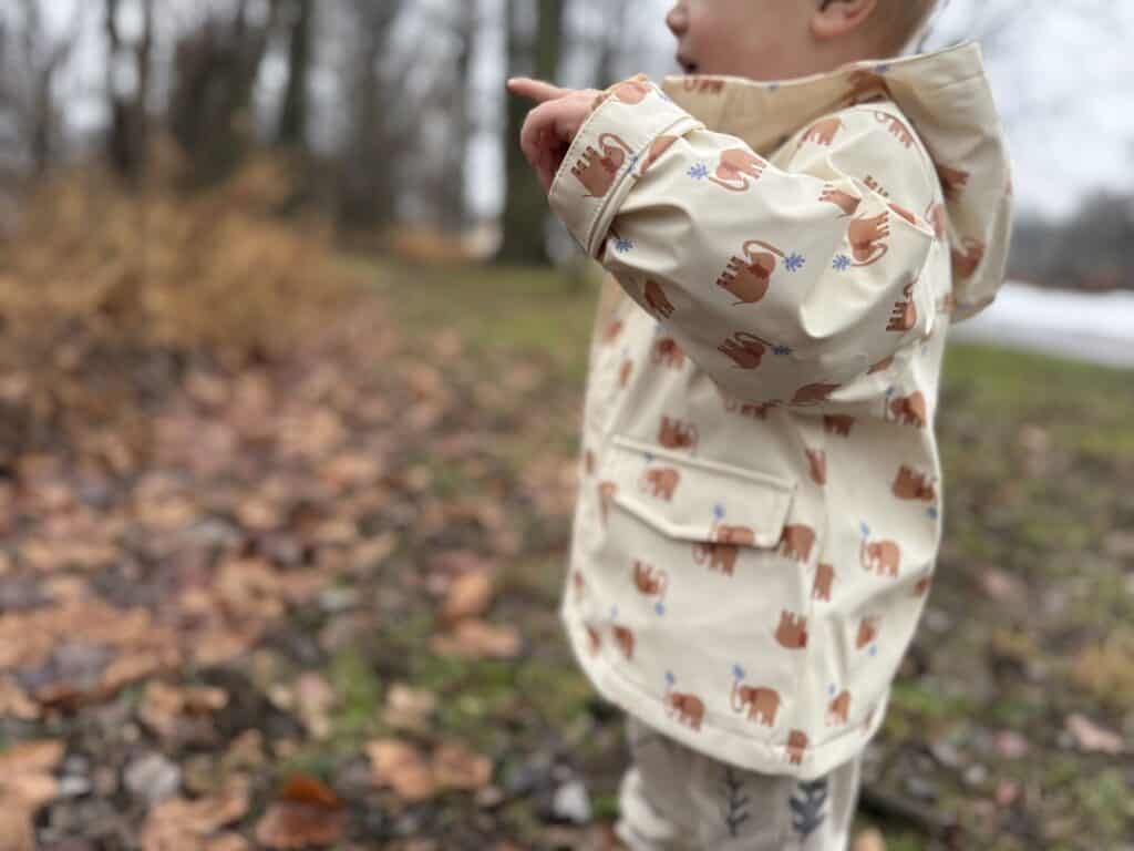 Side view of toddler in Amazon Essentials Toddler Rain Jacket pointing at welt pocket with snap flap