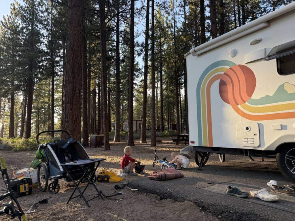 Two young children playing near bikes and a bike trailer at a forest campsite at dusk, with a vintage-style camper van nearby