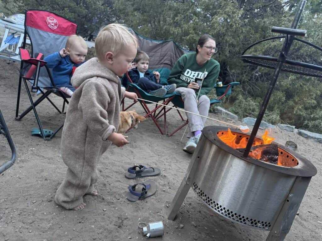 A toddler roasting a marshmallow over a campfire while family members sit nearby