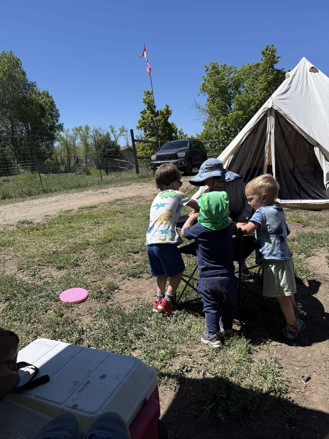Three young children gathered around a camp table at a tent campsite with a vehicle and trees in the background
