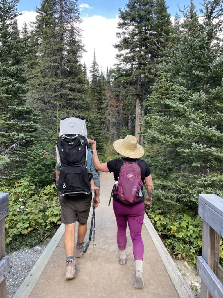 Two hikers with backpacks, trekking poles, and a child carrier walking across a wooden boardwalk in an evergreen forest
