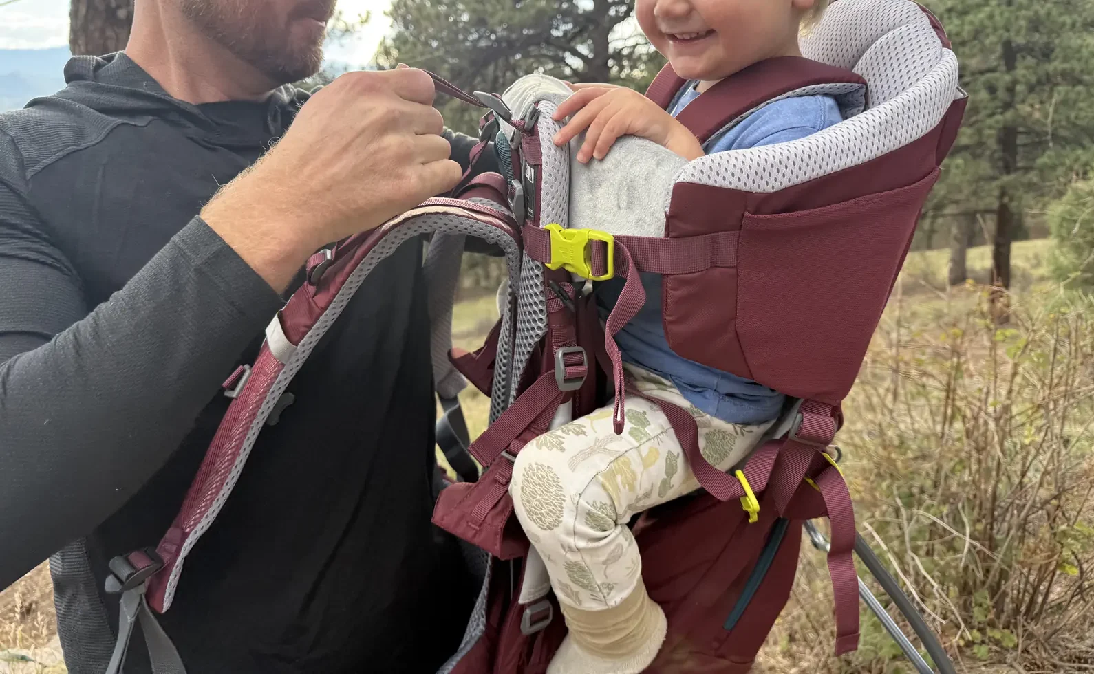 Dad buckling a smiling toddler into the Deuter Kid Comfort child carrier on a pine-covered hillside