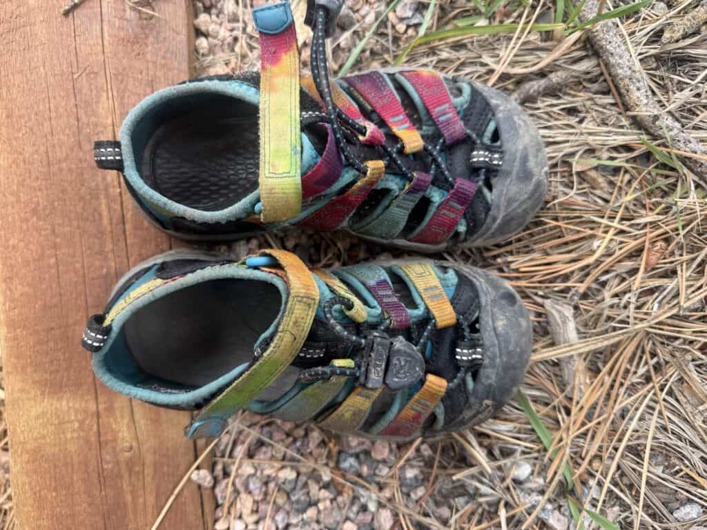 Pair of tie-dye KEEN Newport H2 kids sandals with scuffed toe caps on a wooden step