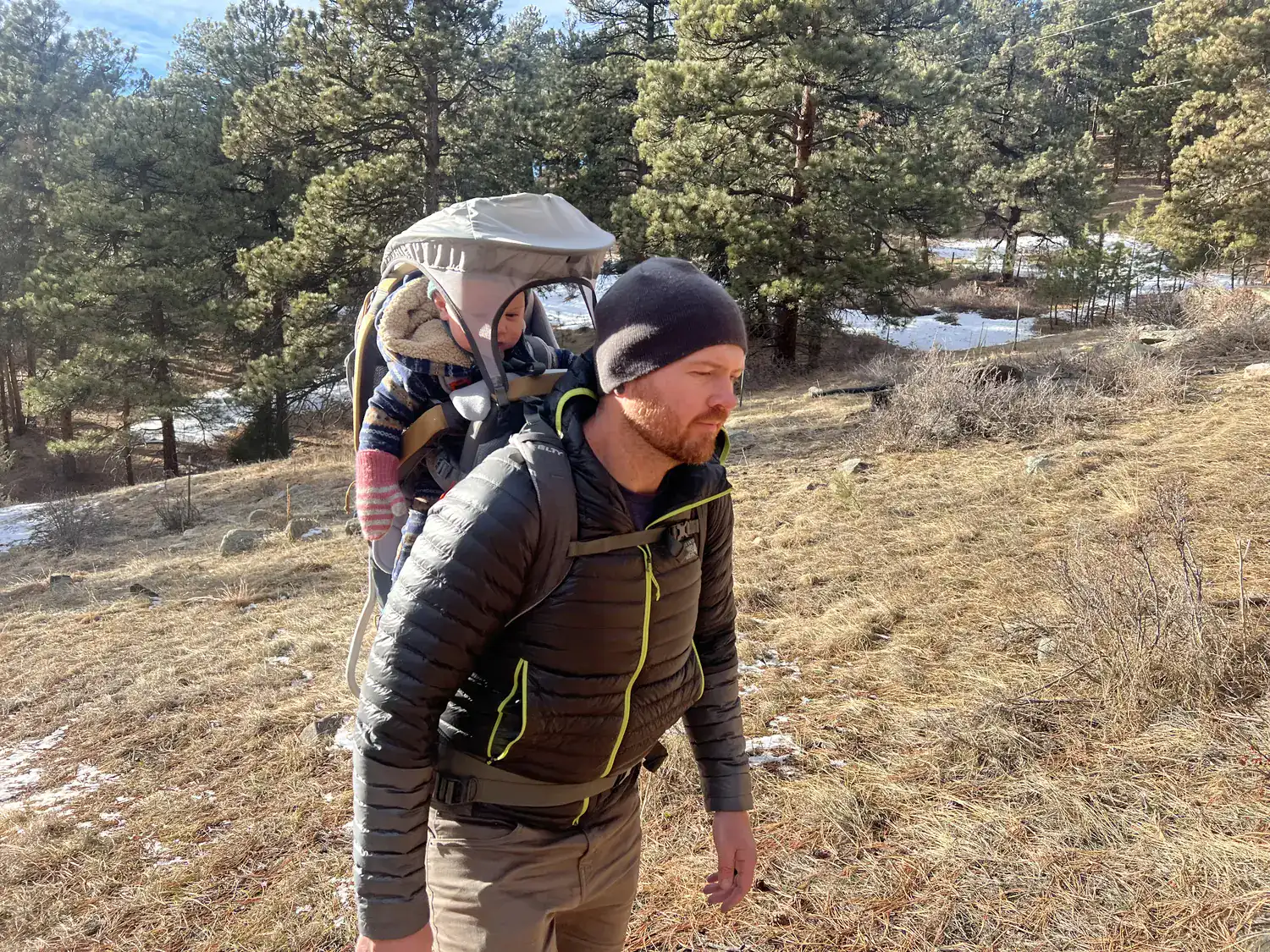 Dad hiking a sunny winter trail with a bundled toddler in the Kelty Journey PerfectFIT Elite child carrier