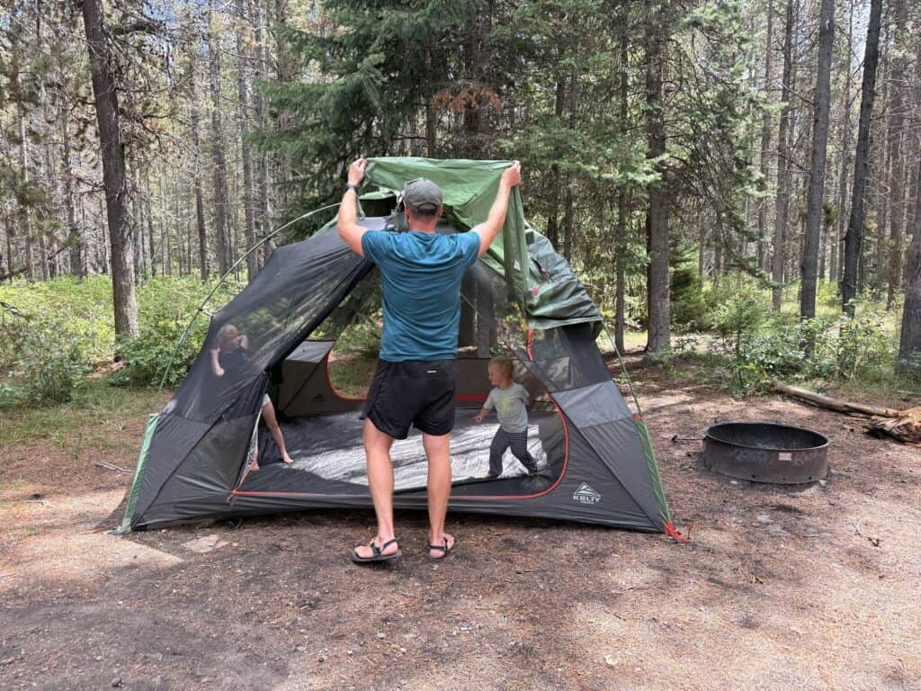 Adult pulling the green rainfly over the Kelty Wireless 6 with a child inside the tent
