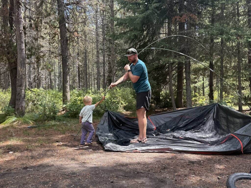 Adult threading a pole on the Kelty Wireless 6 while a young child watches at a forest campsite