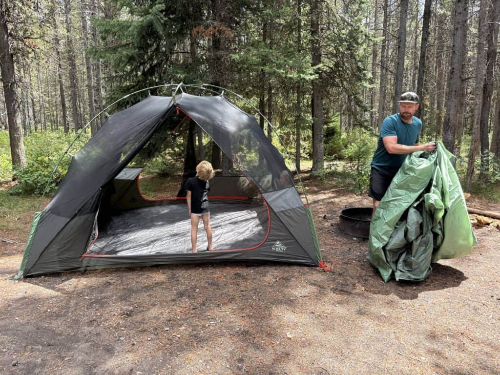 Kelty Wireless 6 tent body pitched with a child inside while an adult unpacks the green rainfly