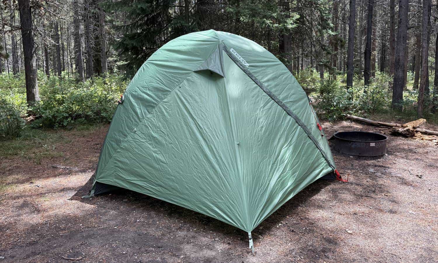 Kelty Wireless 6 tent with its green rainfly pitched at a forest campsite next to a fire ring