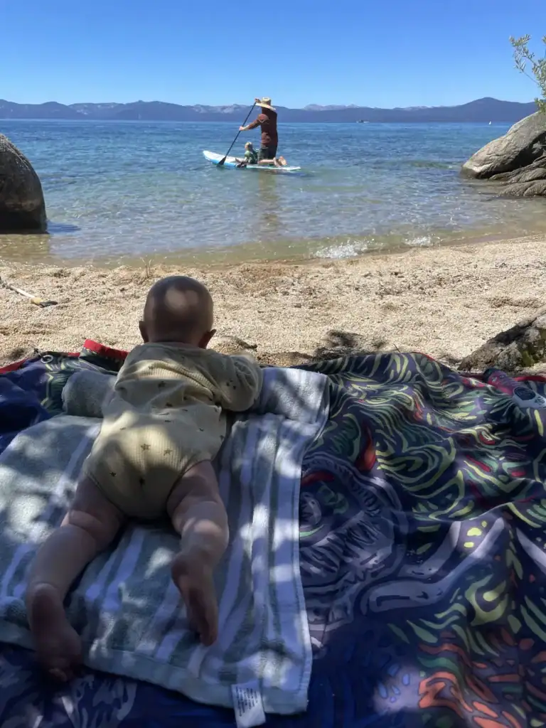 A baby lies on a patterned beach blanket facing turquoise water while an adult and child paddleboard offshore on Lake Tahoe's East Shore.