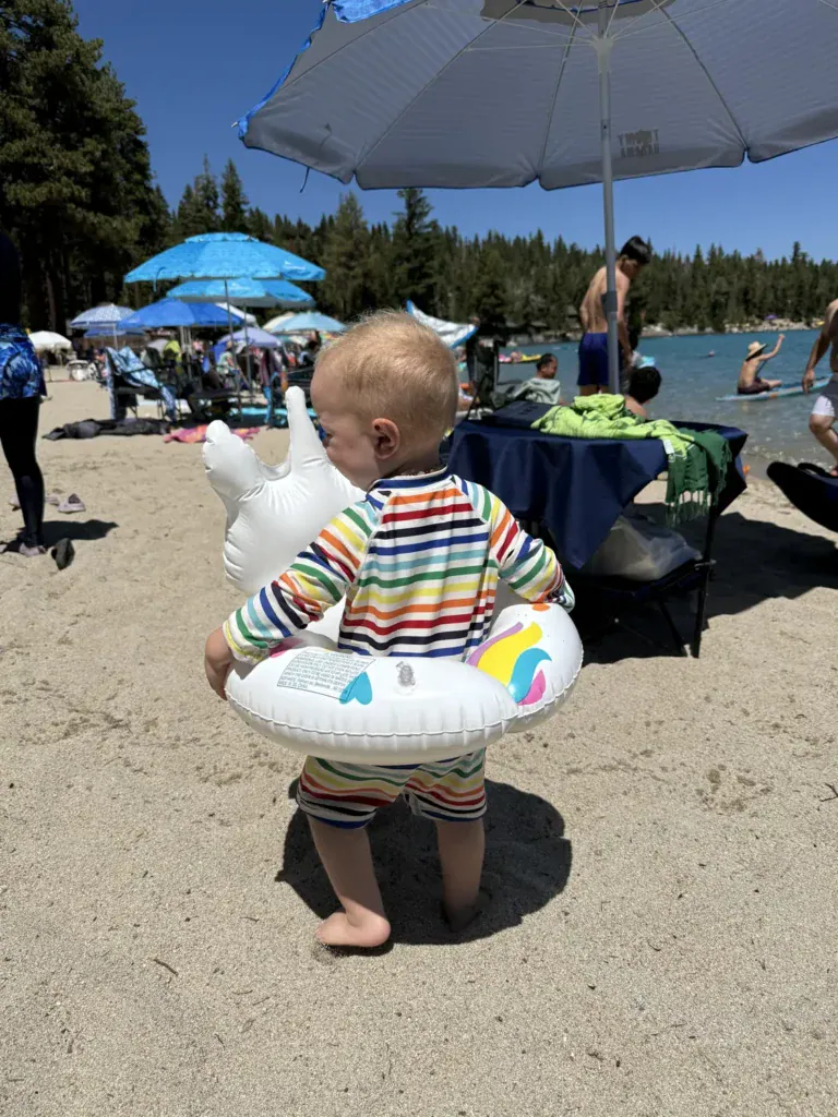 A baby in a striped sun suit holds an inflatable swim float on a busy Lake Tahoe beach full of umbrellas.