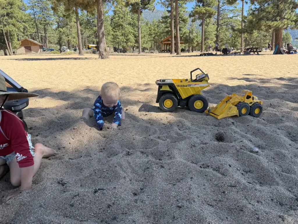 A baby digs in sand next to yellow toy dump trucks under pines near Lake Tahoe.