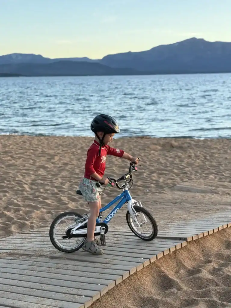 A young kid in a red shirt and helmet rides a balance bike along a wooden boardwalk on a Lake Tahoe beach in evening light.