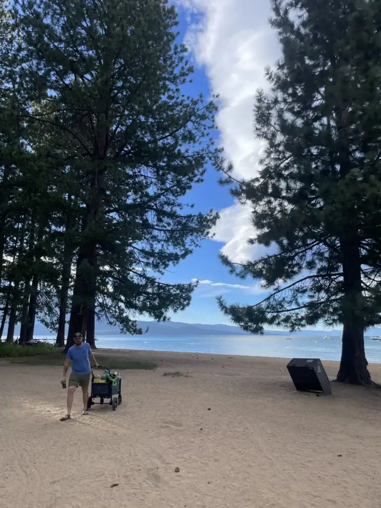 A parent pulls a beach wagon across sand under tall pines toward Lake Tahoe.