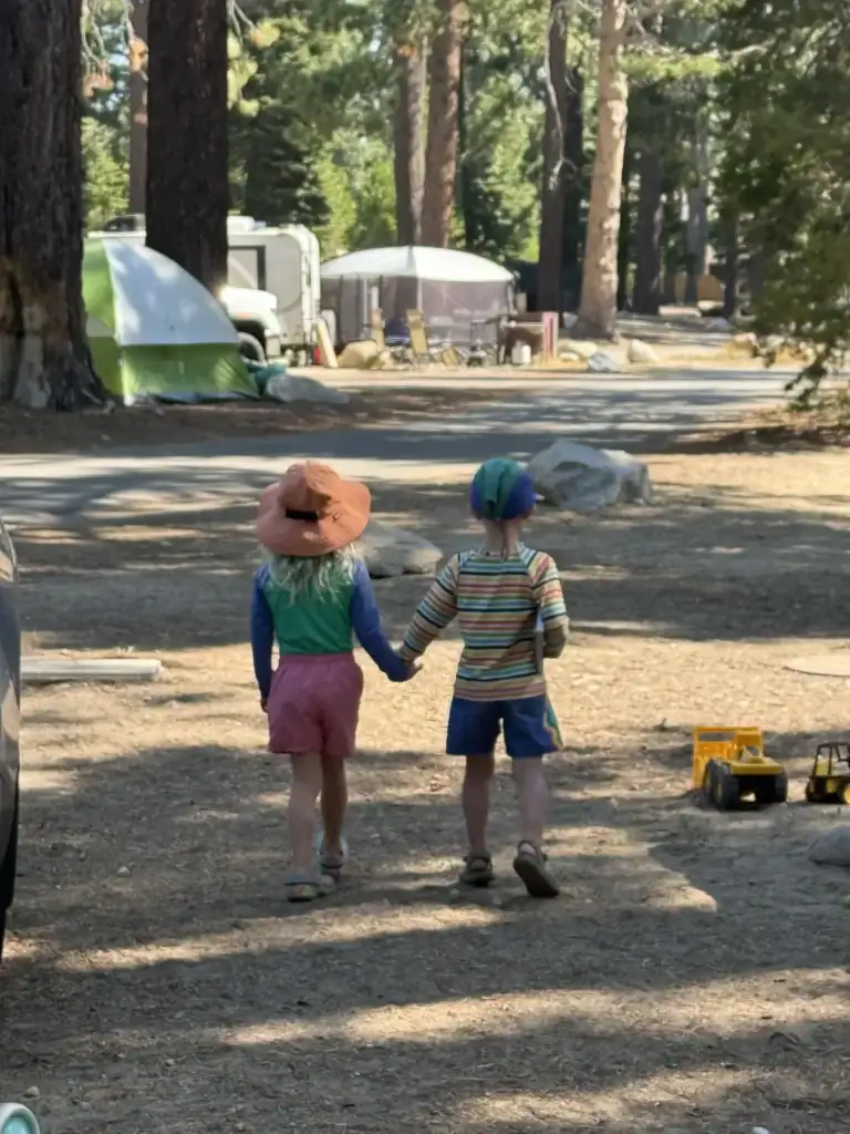 Two young kids in sun hats walk hand in hand from behind through a pine-shaded Lake Tahoe campground loop.