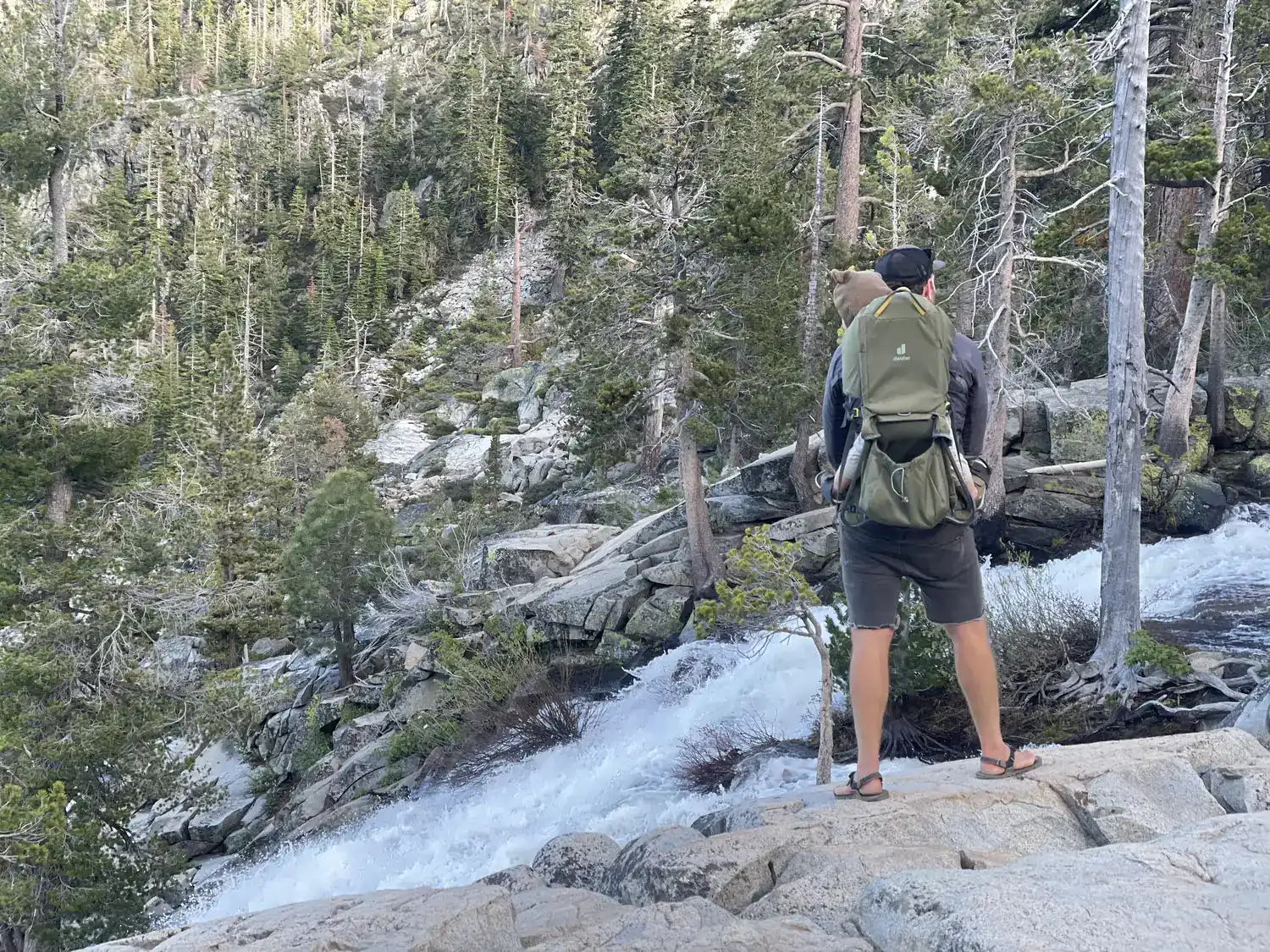 An adult wearing a child hiking carrier stands on granite slabs above the rushing top of Cascade Falls near Emerald Bay, Lake Tahoe.