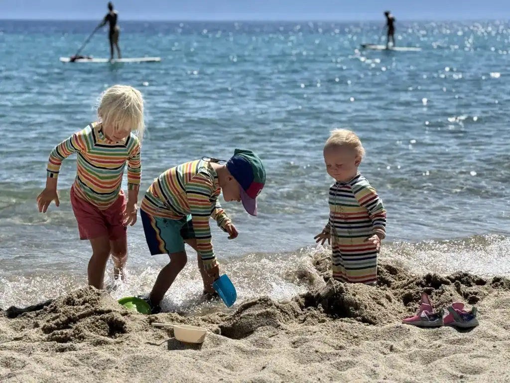 Three kids in sun shirts dig in the sand at the edge of Lake Tahoe while paddleboarders pass behind them.