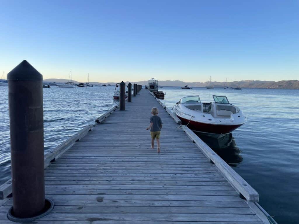 A toddler walks away from the camera down a long wooden dock on Lake Tahoe at dusk, boats moored nearby.