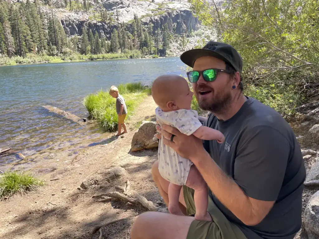 A parent holds a baby on the shore of Eagle Lake while a toddler explores the water's edge below granite cliffs.