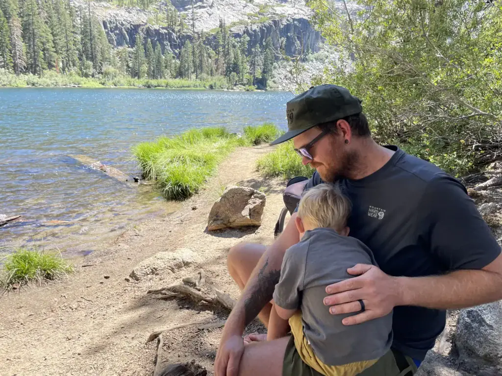 A parent hugs a toddler on the rocky shore of Eagle Lake, with granite cliffs and pines across the water.