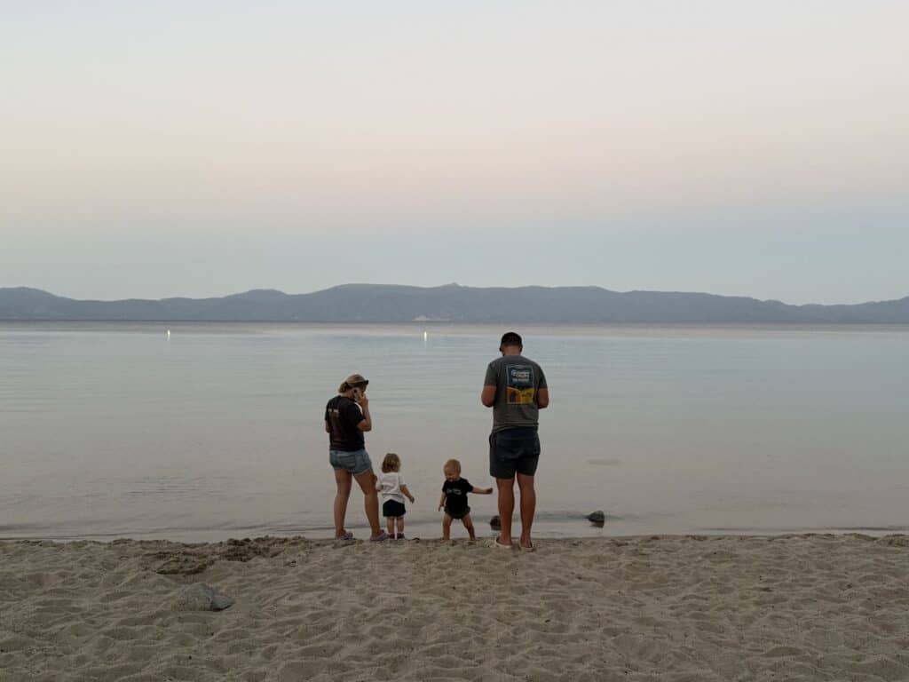 Adults and two small kids stand at the edge of calm Lake Tahoe at dusk with mountains across the water.