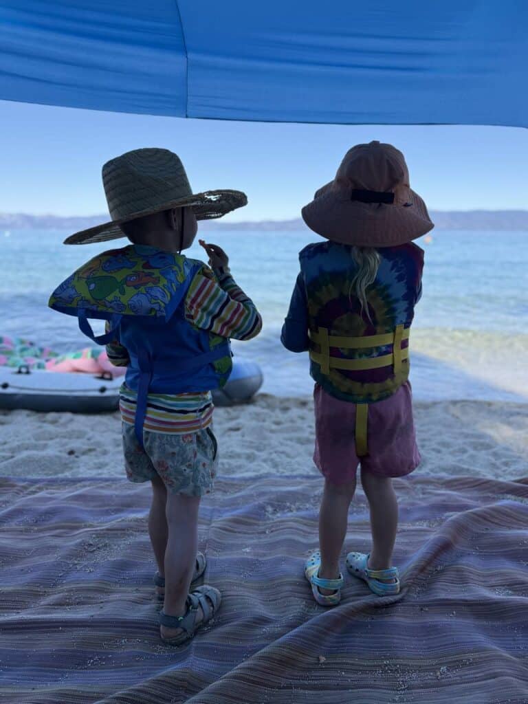Two kids seen from behind in life jackets and wide-brim sun hats stand on a blanket facing Lake Tahoe.