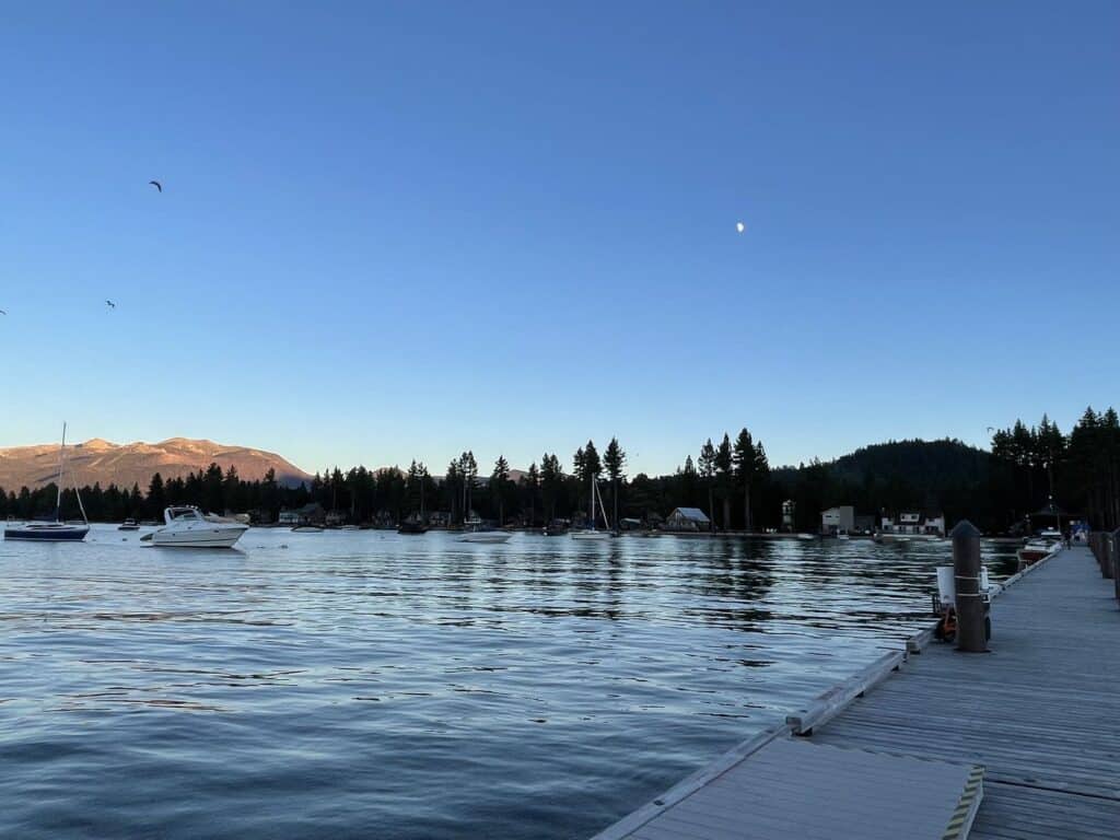 Boats sit in a calm Lake Tahoe marina in evening light beside a wooden dock and pine-covered shoreline.