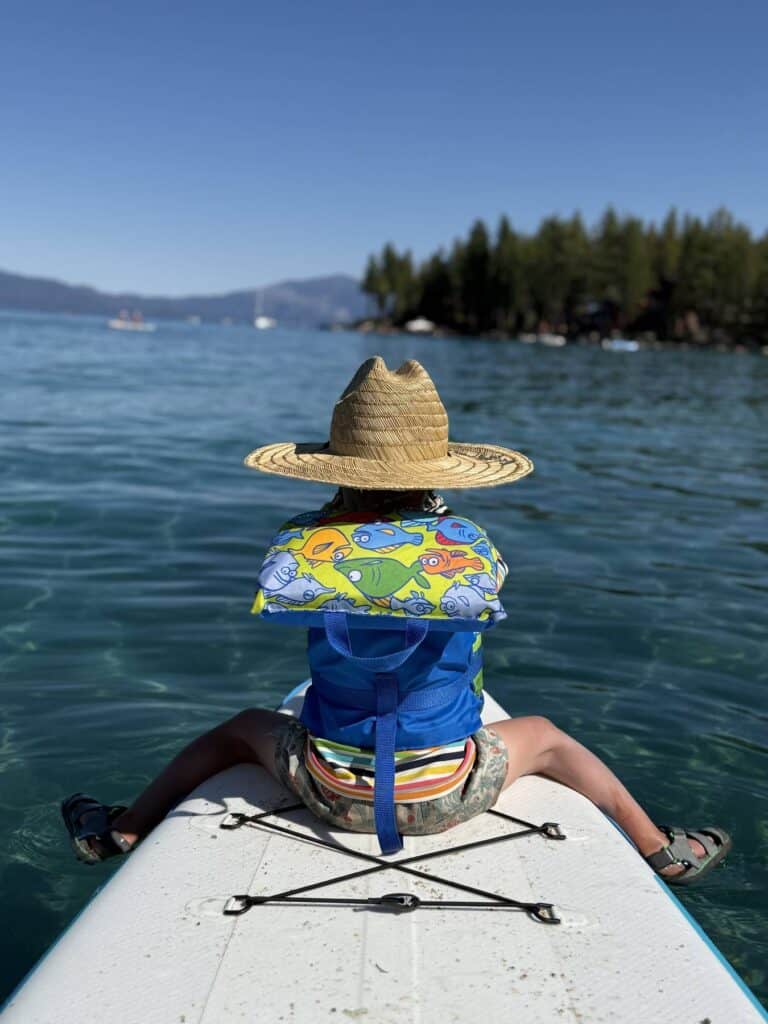 A child in a life jacket and straw sun hat sits on the front of an inflatable paddleboard on clear Lake Tahoe water.
