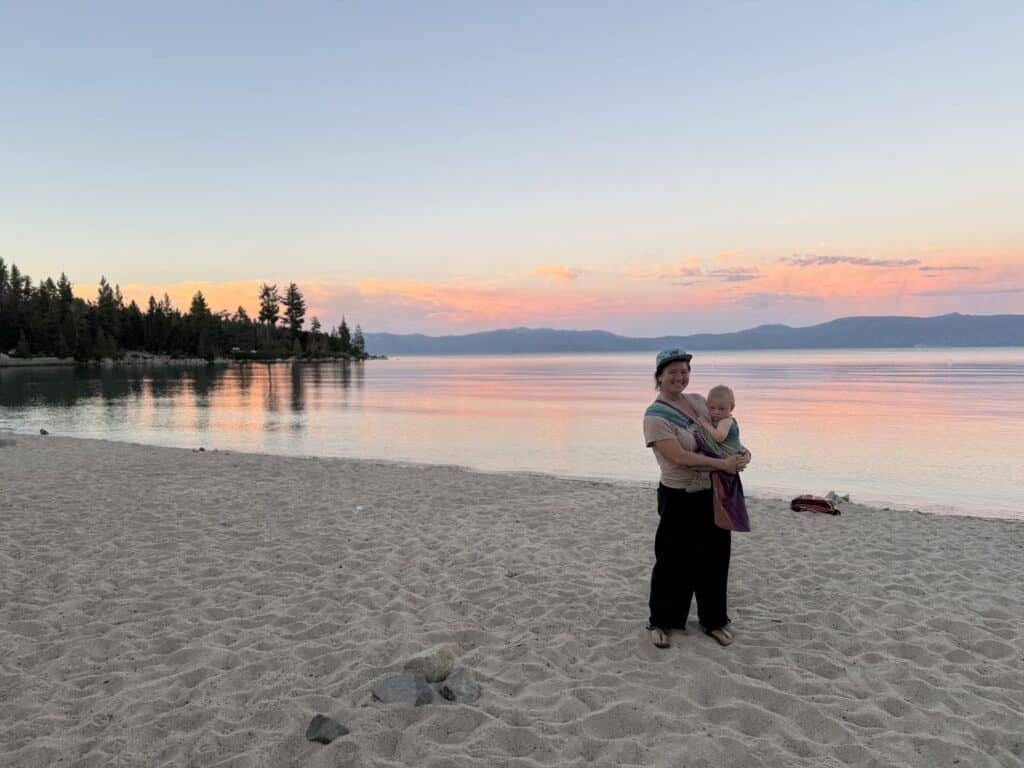 A parent holds a baby on a sandy Lake Tahoe beach under a pink and orange sunset.