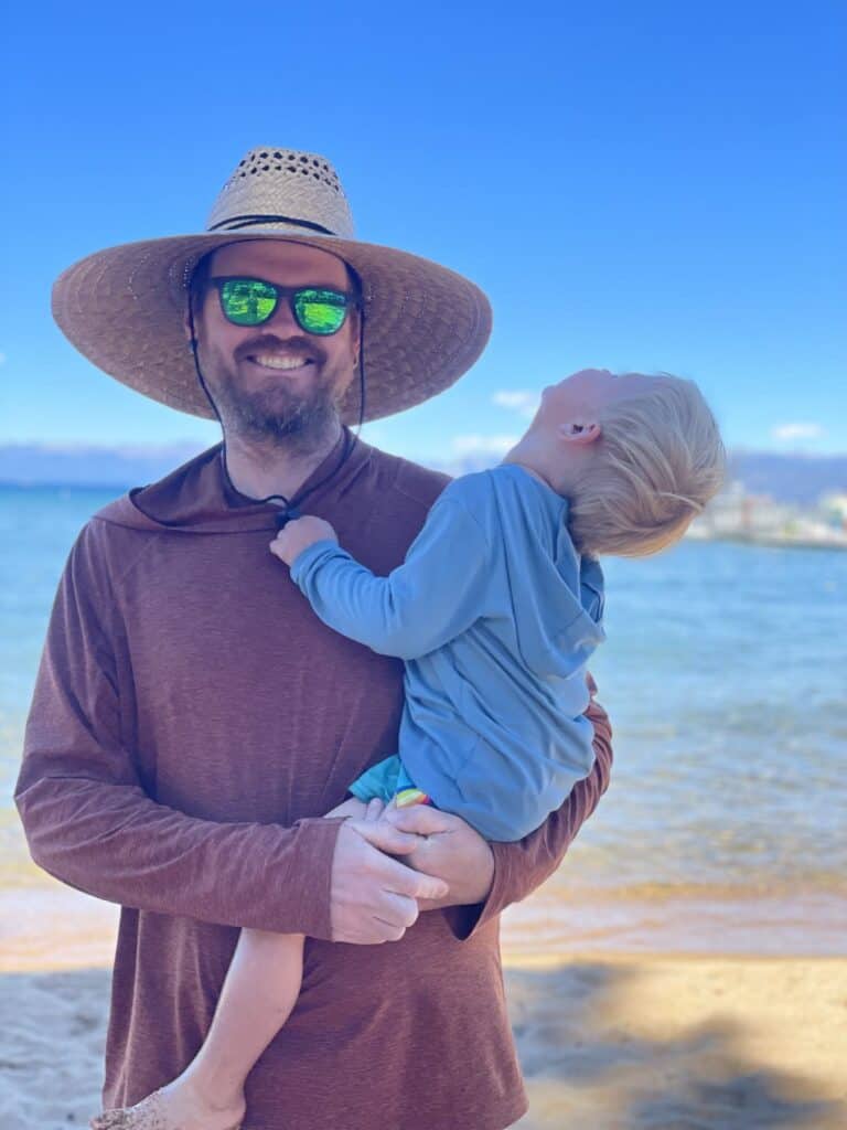 A parent in a wide-brim straw hat holds a toddler in a blue sun shirt on a Lake Tahoe beach.