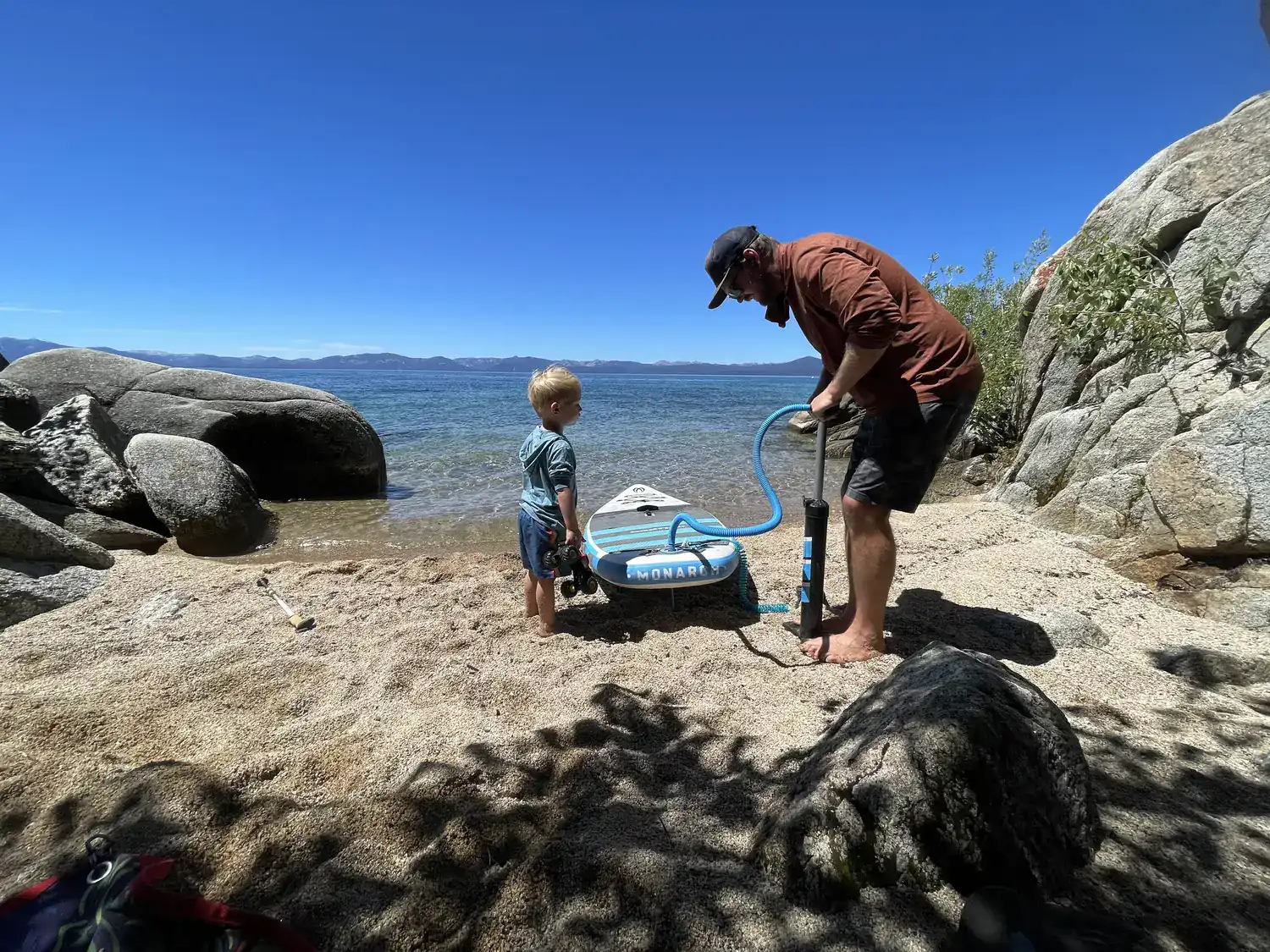A parent inflates a paddleboard with a hand pump while a toddler watches on a granite cove on Lake Tahoe's East Shore.