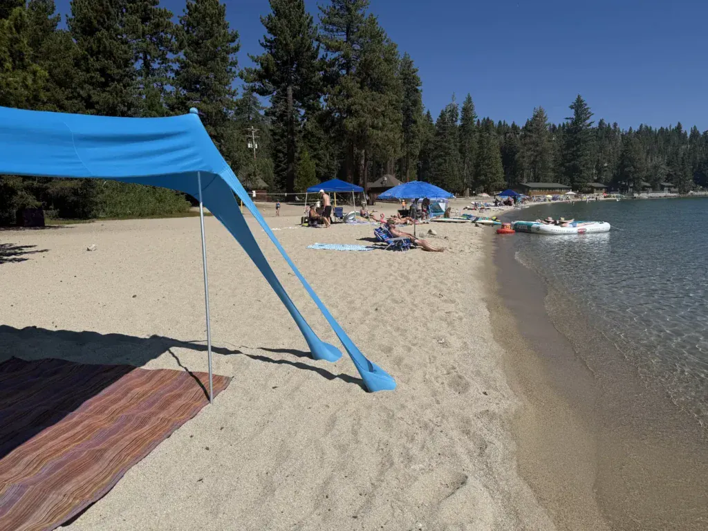 A blue shade canopy is staked in the sand over a striped blanket on a Lake Tahoe beach lined with pines.