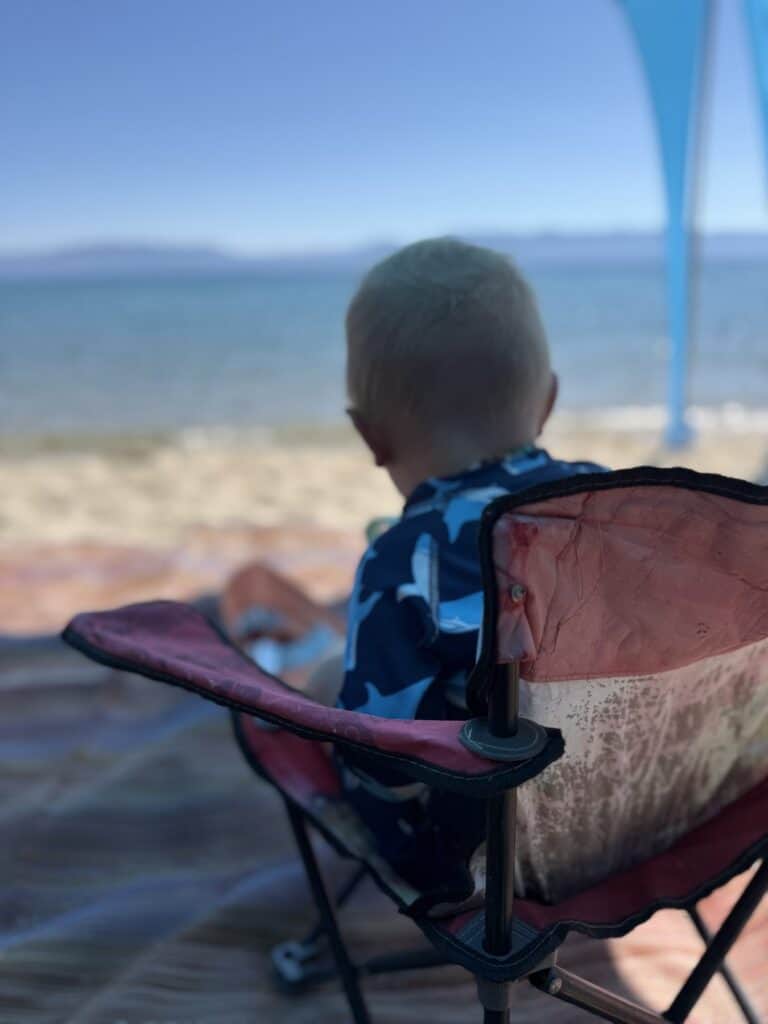A toddler seen from behind sits in a small red camp chair facing blue Lake Tahoe under a beach canopy.