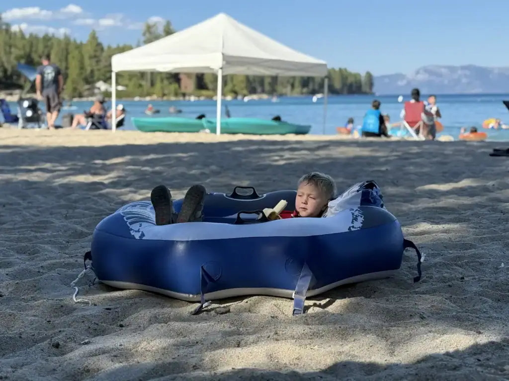 A toddler relaxes inside a blue inflatable float on the sand at a busy Lake Tahoe beach.