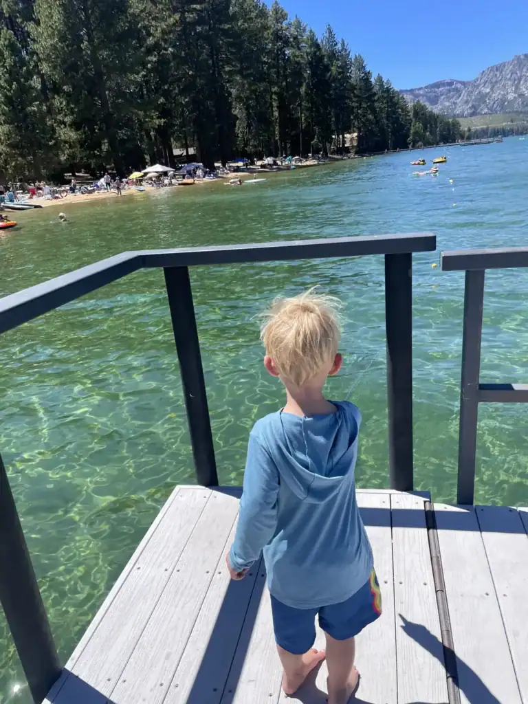 A toddler in a blue sun hoodie stands on a pier looking over clear green Lake Tahoe water toward a busy beach.