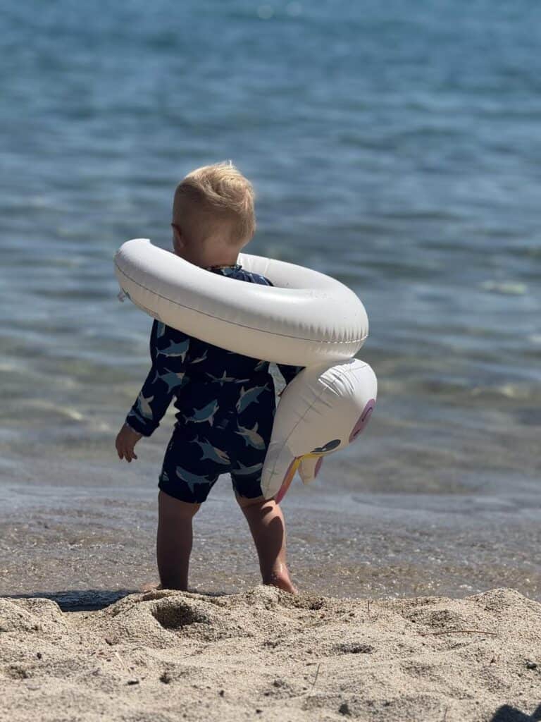 A toddler in a sun-protective swimsuit wearing an inflatable swim float stands at the edge of Lake Tahoe.