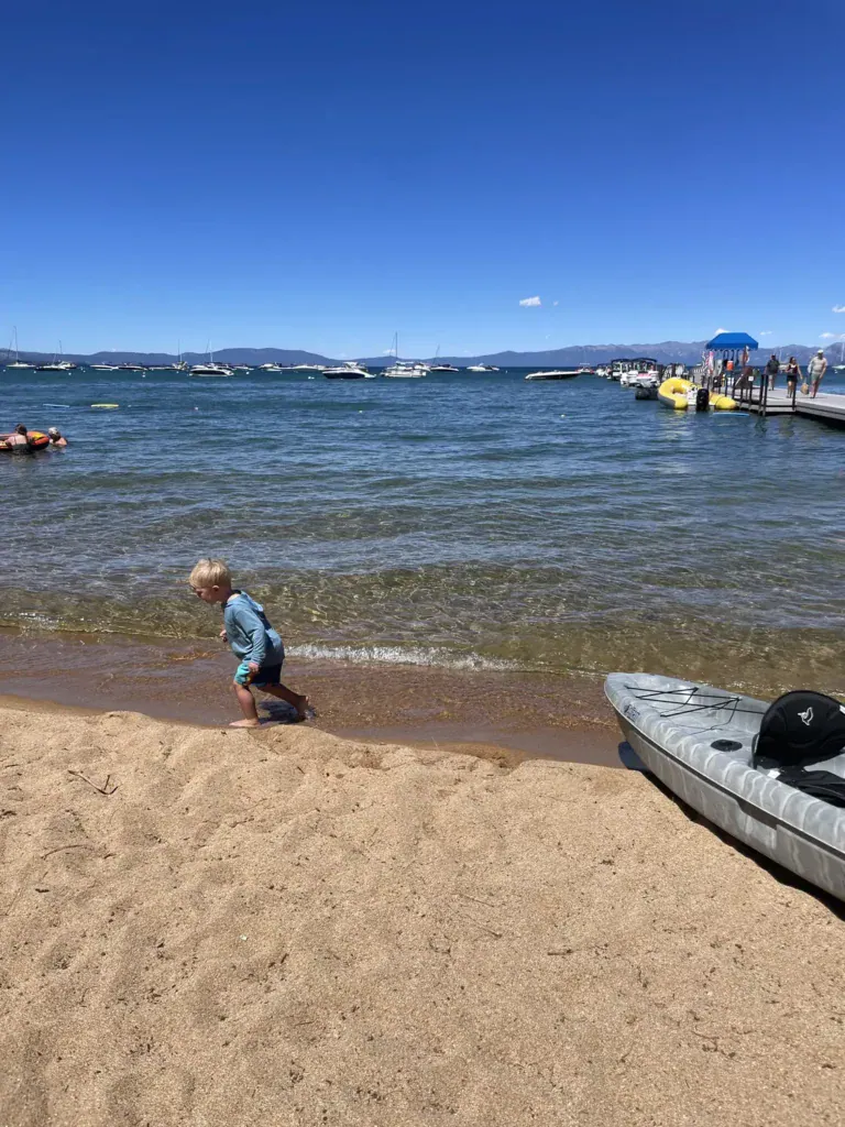 A toddler wades at the edge of Lake Tahoe on a sandy beach with boats and a pier in the background.