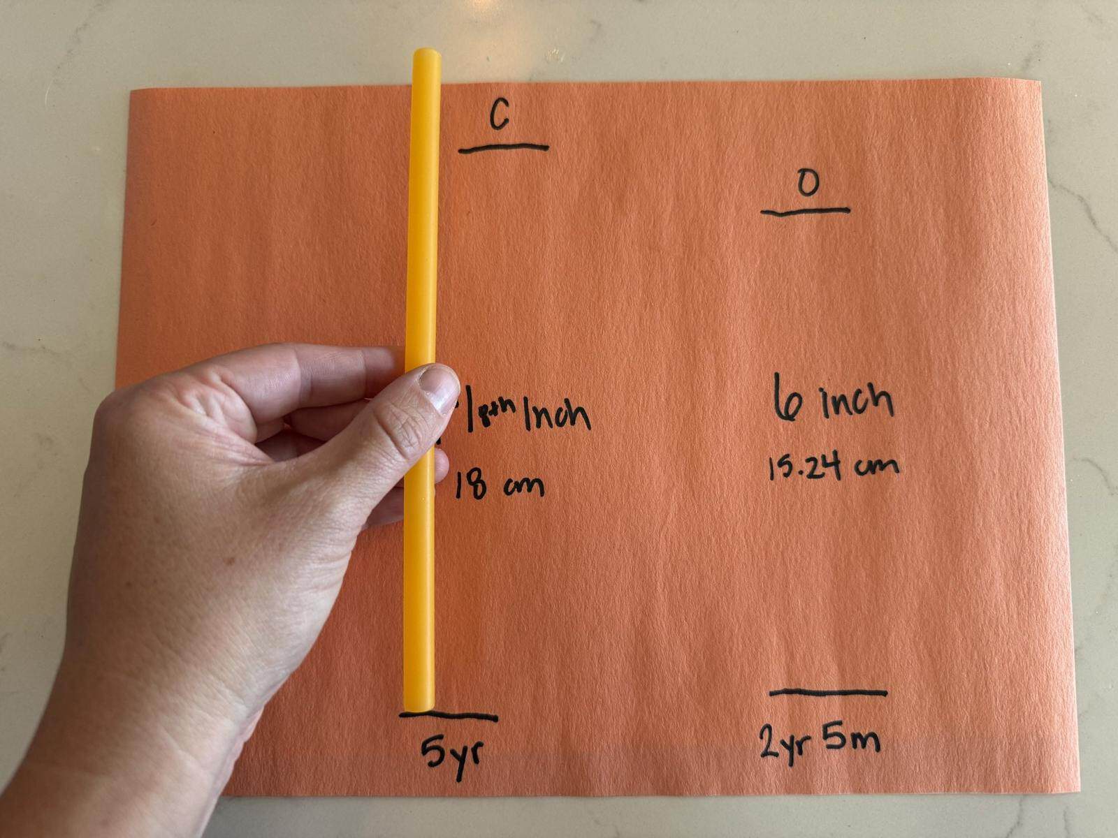 Hand holding a yellow straw lined up with a child's heel and toe lines on orange paper, with measurements of 7 1/8 inch, 18 cm and 6 inch, 15.24 cm written beside them