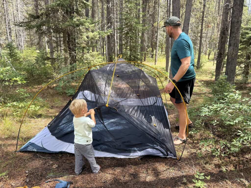 Toddler standing beside the Mountain Hardwear Mineral King 3 tent body as an adult sets it up in a forest